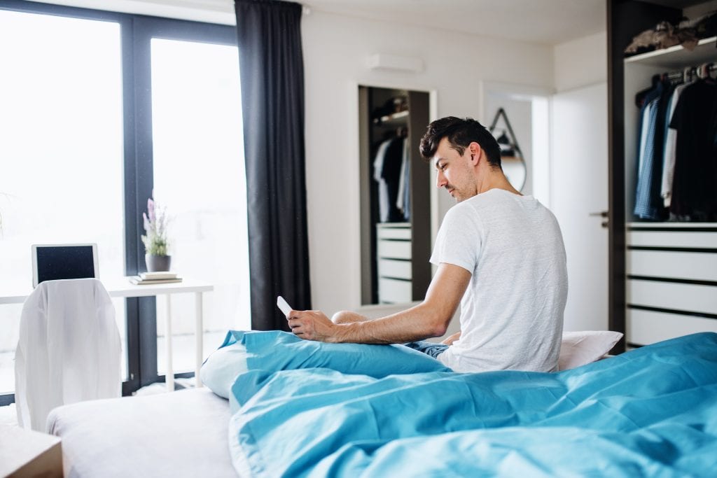 Young man with smartphone in bed at home, text messaging.