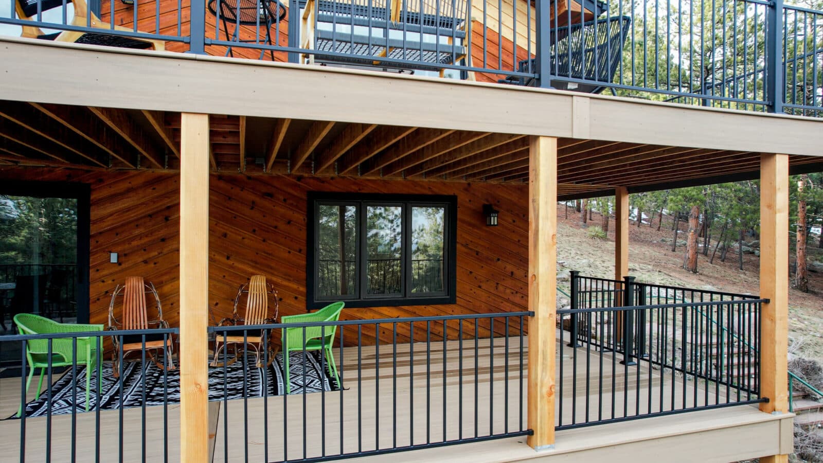 A two-level composite deck with black metal railings, wood support posts, and a covered lower patio feature outdoor seating.