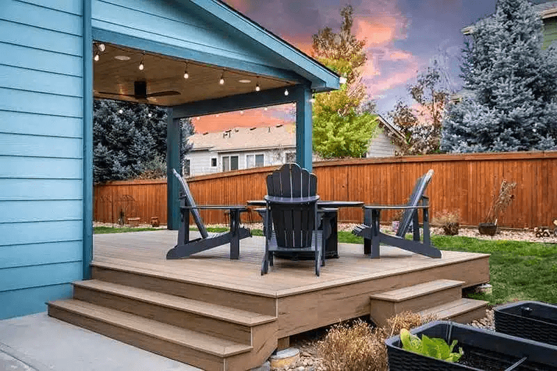 Composite deck with a covered pergola, string lights, Adirondack chairs, and backyard landscaping at sunset.