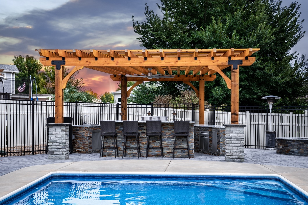 Poolside outdoor kitchen with a wooden pergola, stone bar, and built-in appliances, perfect for entertaining.
