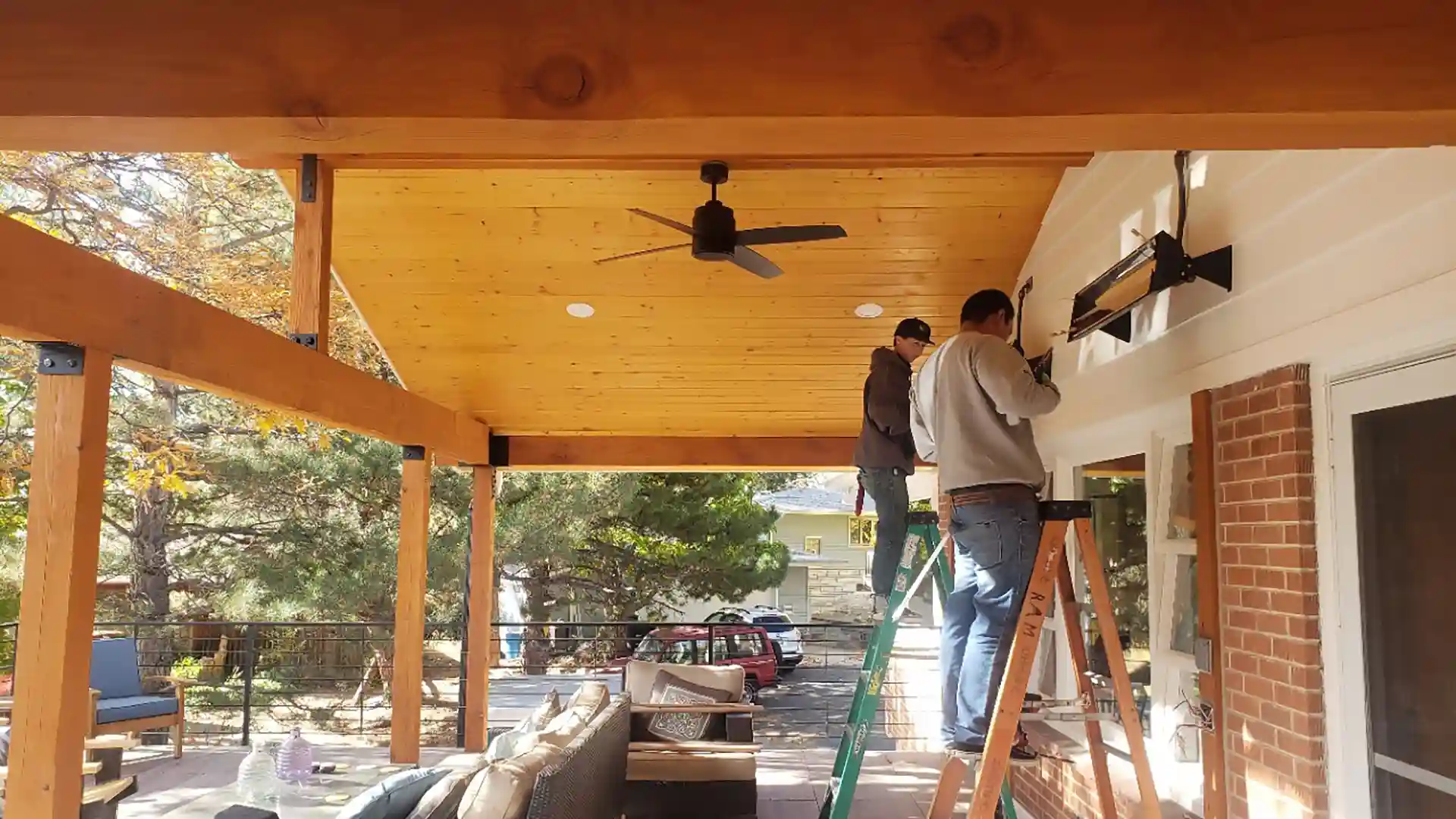 Workers installing a heater under a covered deck with wooden beams and a ceiling fan.