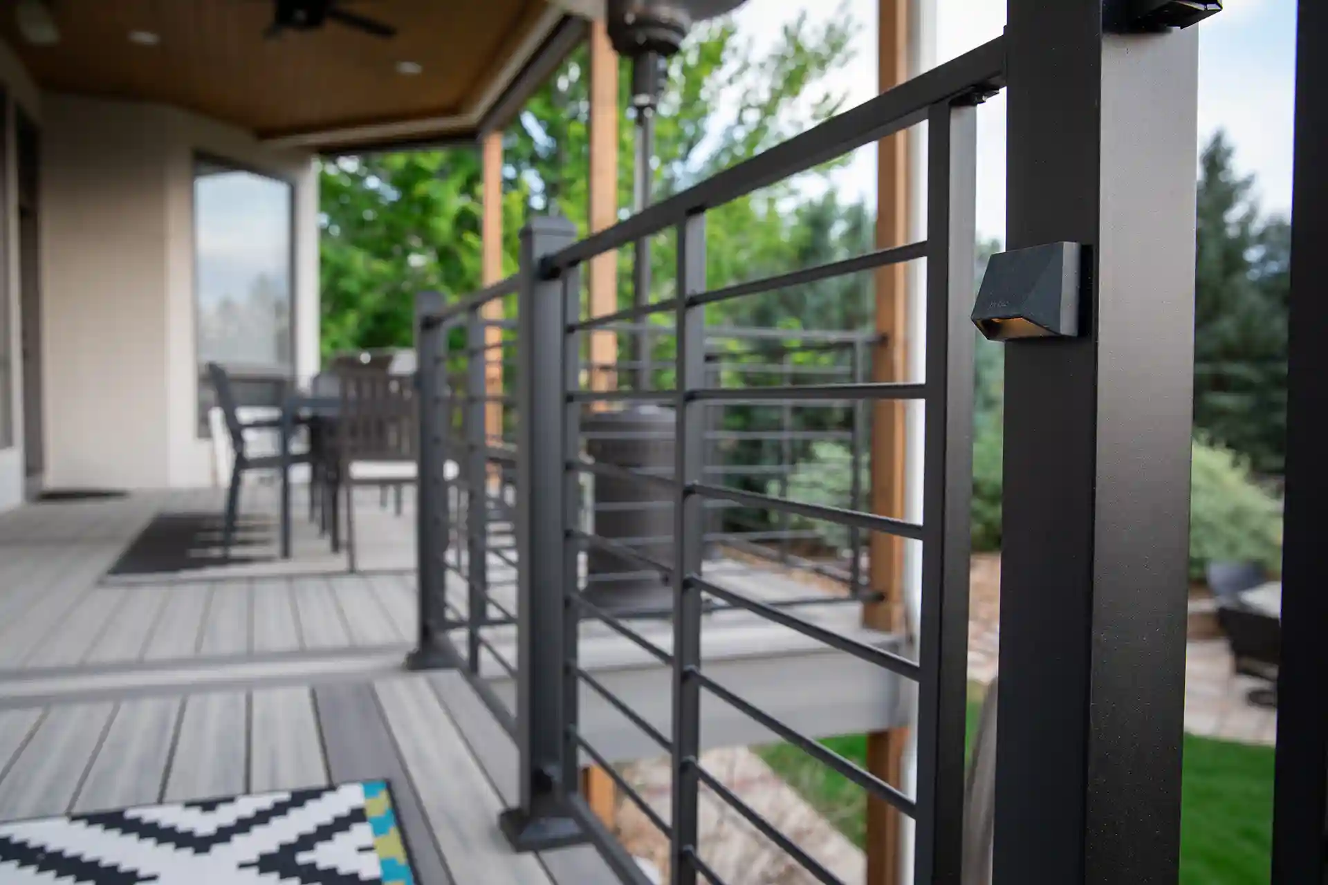 A black aluminum deck railing with horizontal cables on a wooden deck is surrounded by greenery.