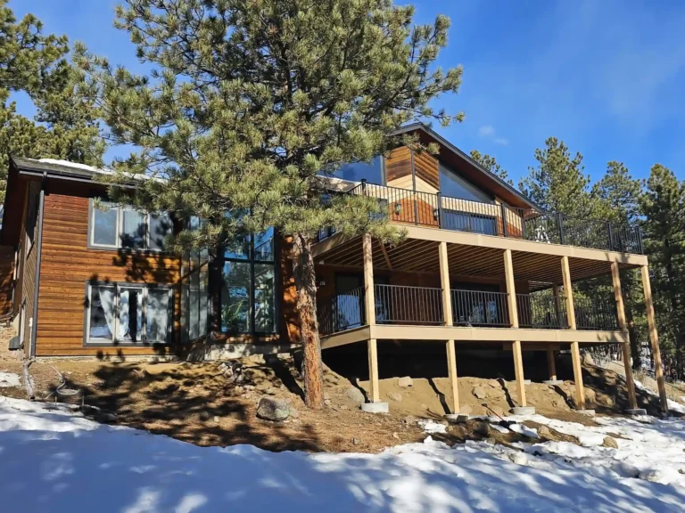 Wooden house with a large two-level deck in a snowy, pine-filled landscape under a clear blue sky.