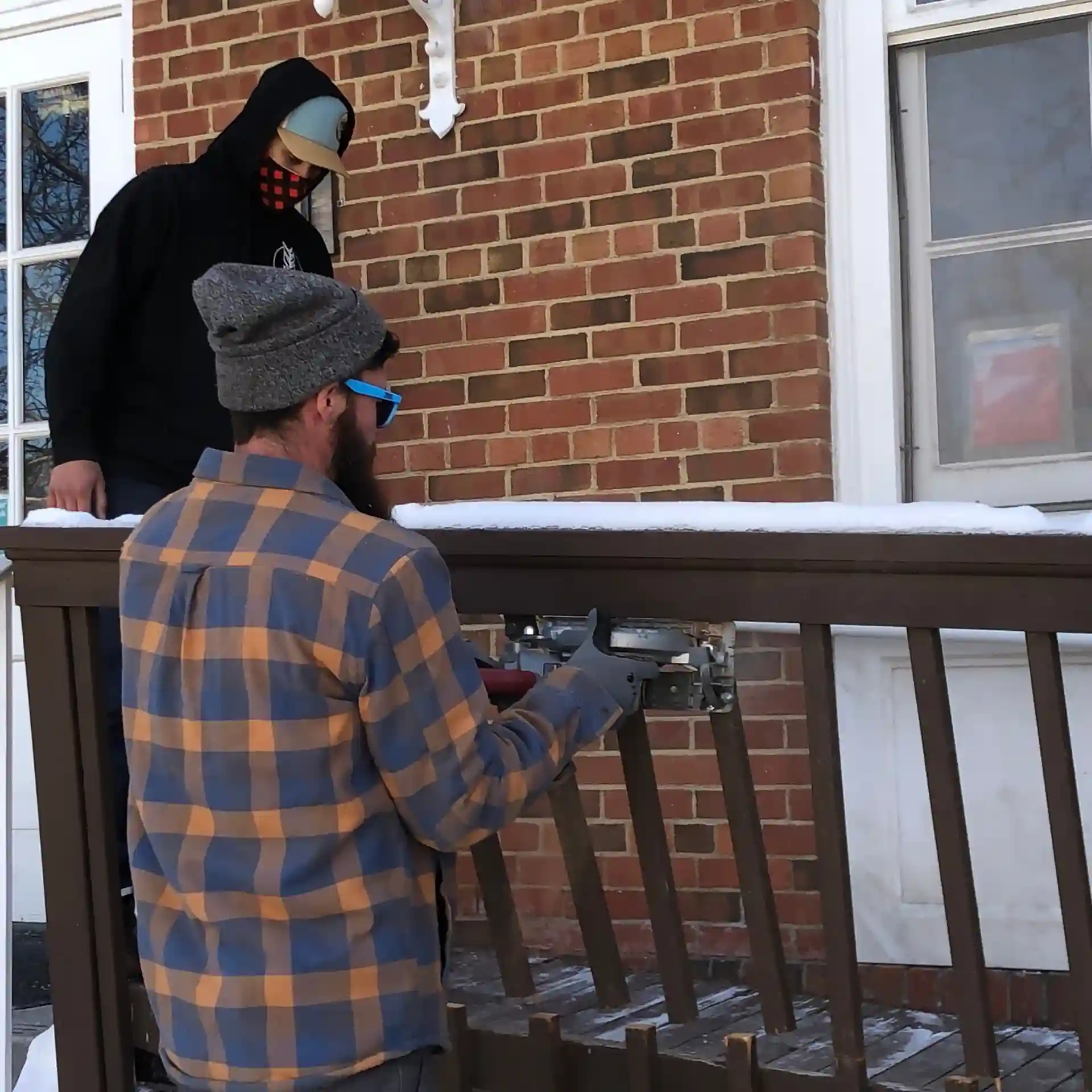 Two men working on repairing a wooden deck railing outside a brick house in winter, with snow visible on the railing. 