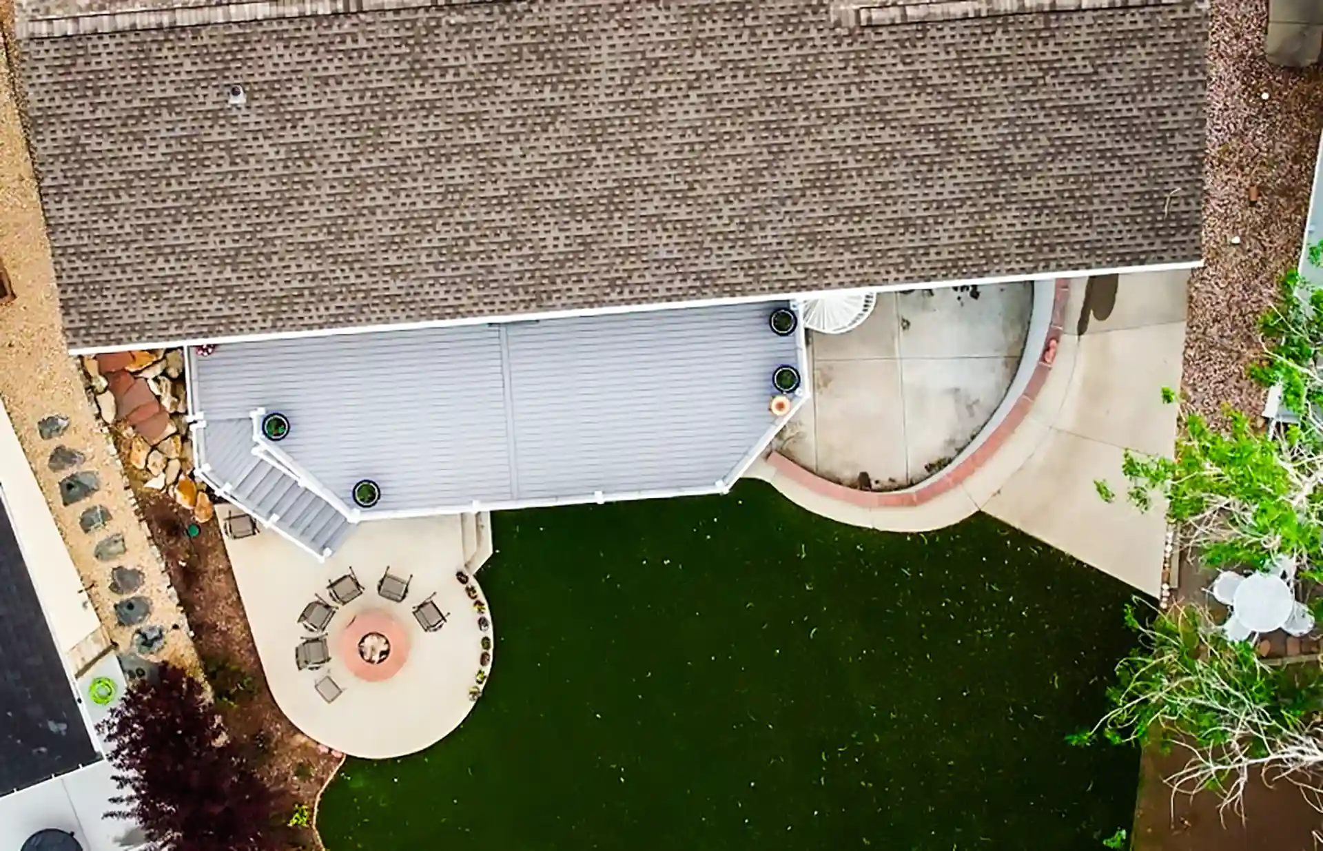Aerial view of a backyard featuring a house with a covered deck, a circular patio with a fire pit surrounded by chairs, and a green lawn bordered by landscaping. 
