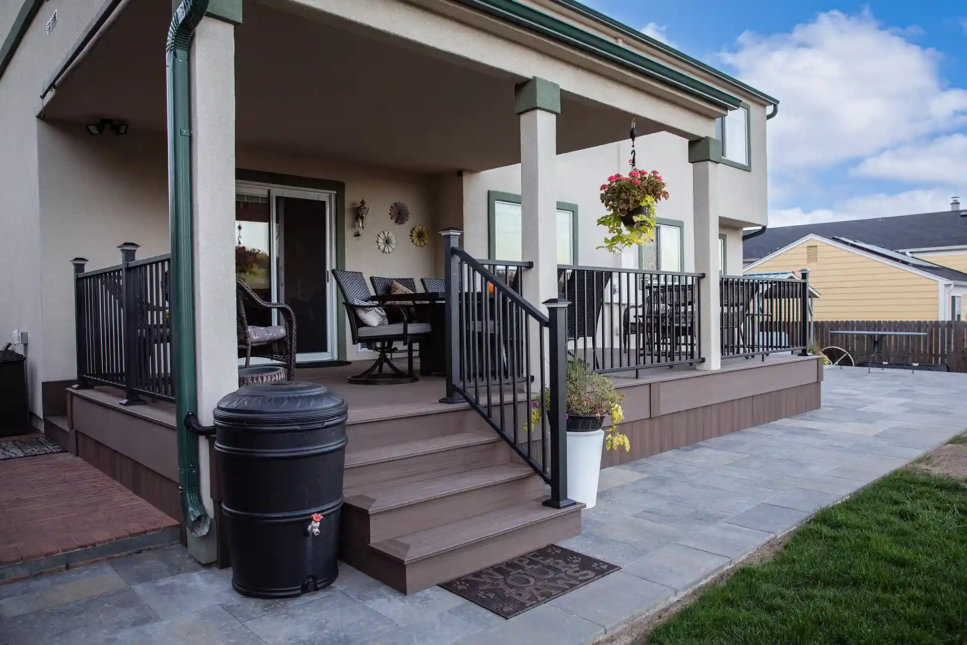 A well-maintained outdoor deck with black metal railings, a set of patio chairs, and a table. The deck is adorned with hanging plants and features a trash bin and a potted plant on the steps.
