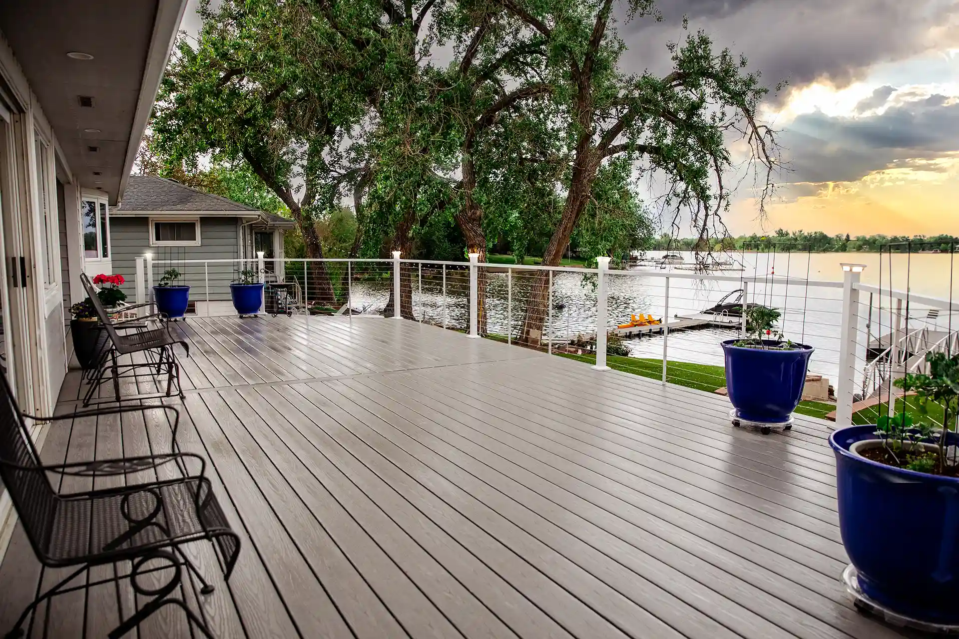 A deck overlooking a lake, featuring white cable railings and large blue planters. The deck features two metal chairs, offering a view that includes trees, lush green grass, and a dock with boats in the distance.
