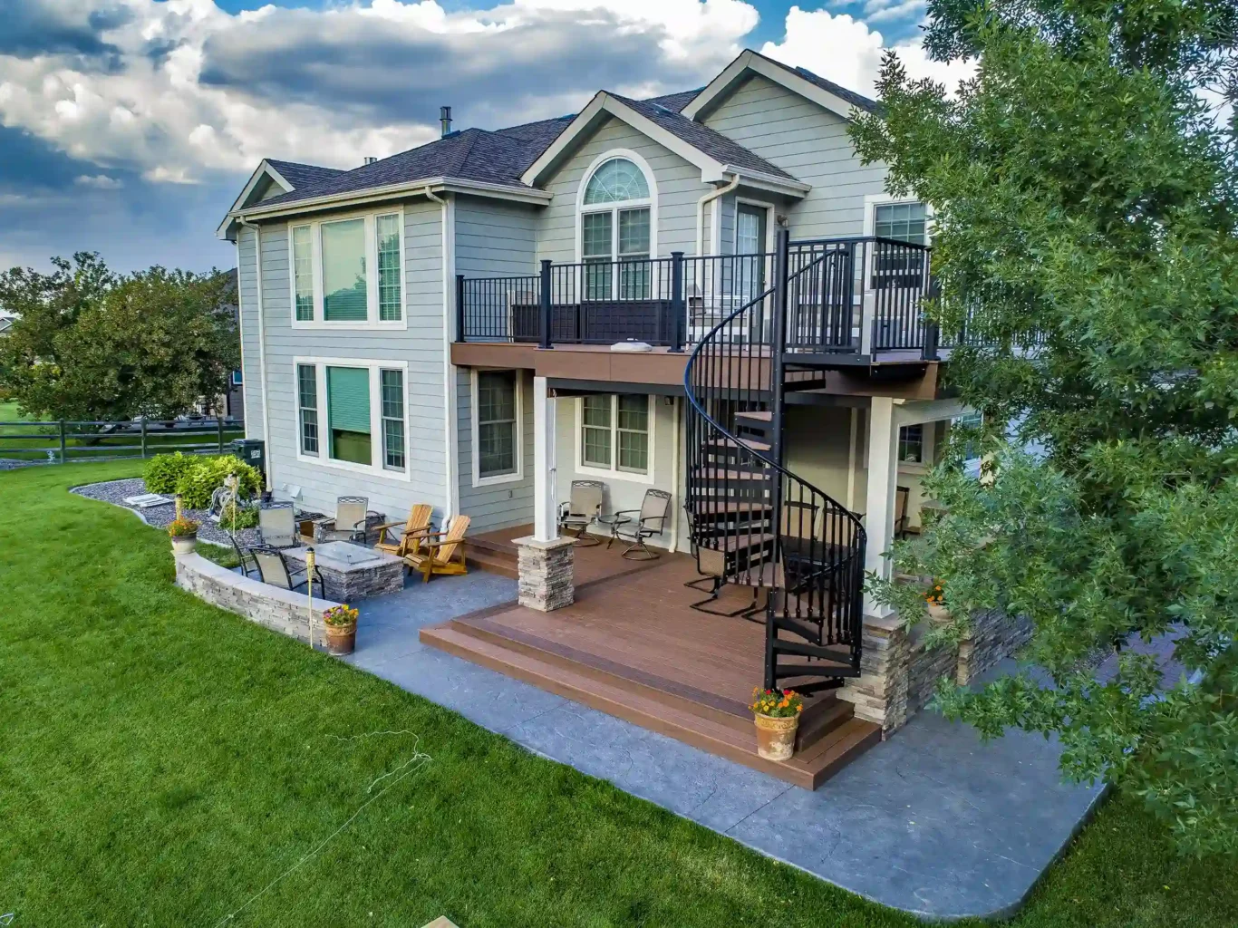 A house with a deck and a spiral staircase leading to an upper level. The deck is surrounded by landscaping with stone planters and a grassy yard.