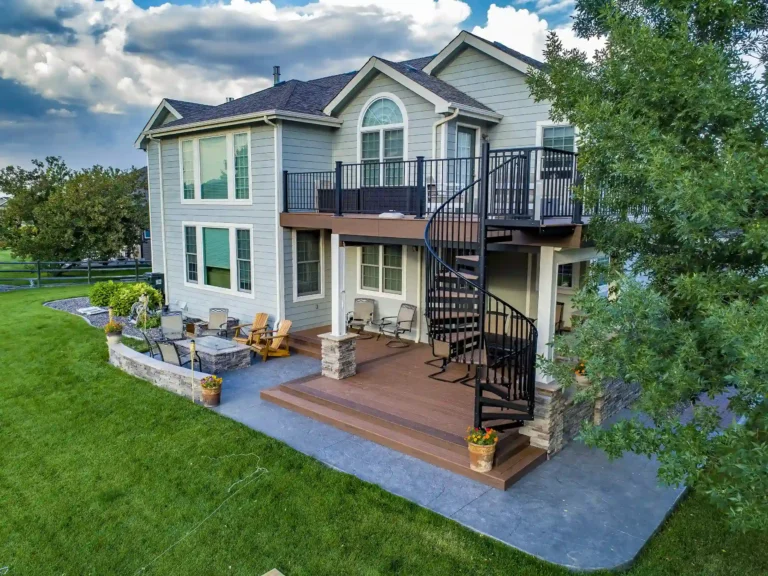A house with a deck and a spiral staircase leading to an upper level. The deck is surrounded by landscaping with stone planters and a grassy yard.