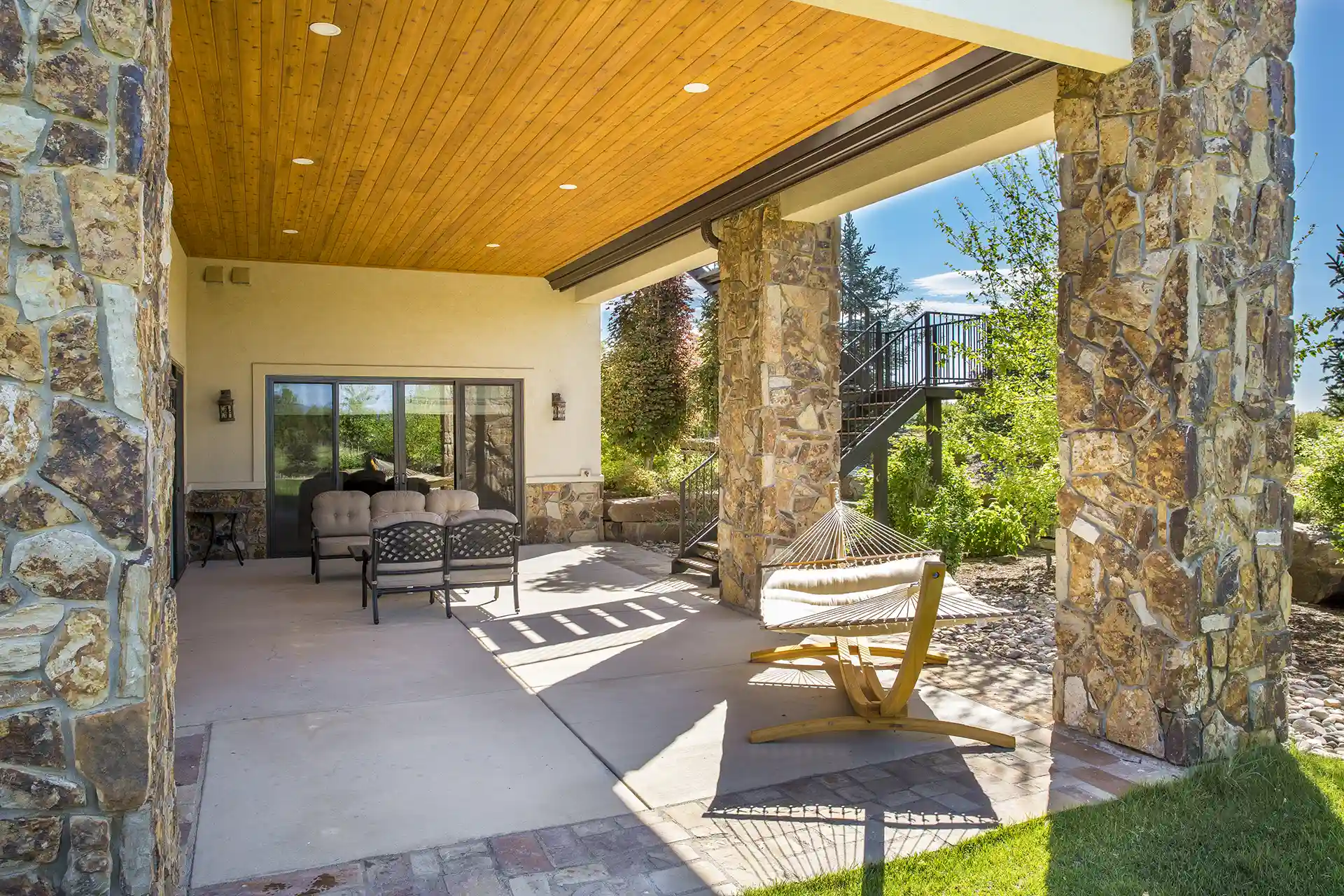 A covered outdoor space with stone pillars, a wooden ceiling, and a hammock. There are two chairs and a sliding glass door leading into the house.
