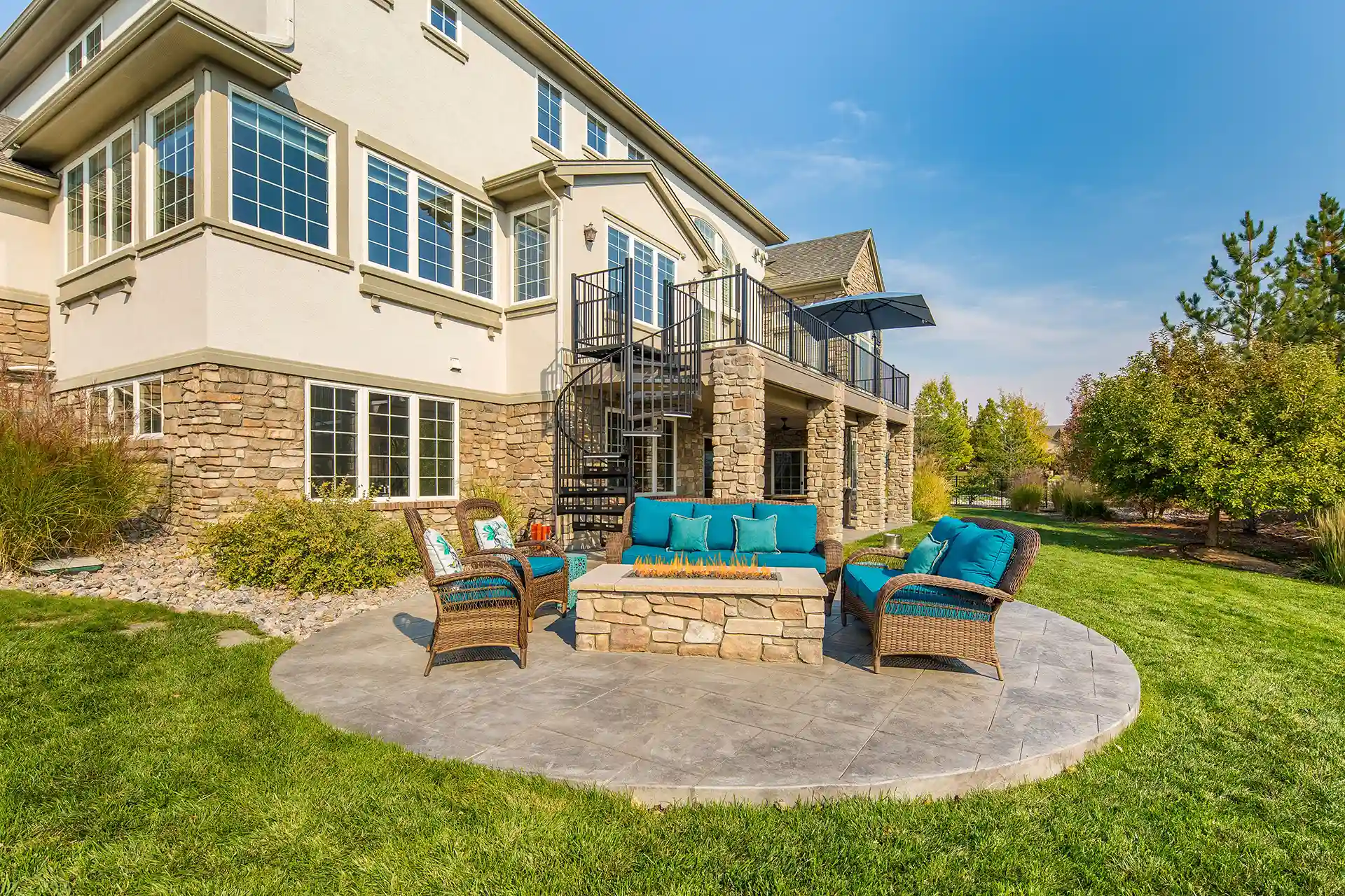 An outdoor seating area with a stone firepit surrounded by chairs with blue cushions. A staircase leads up to the second level of the house.
