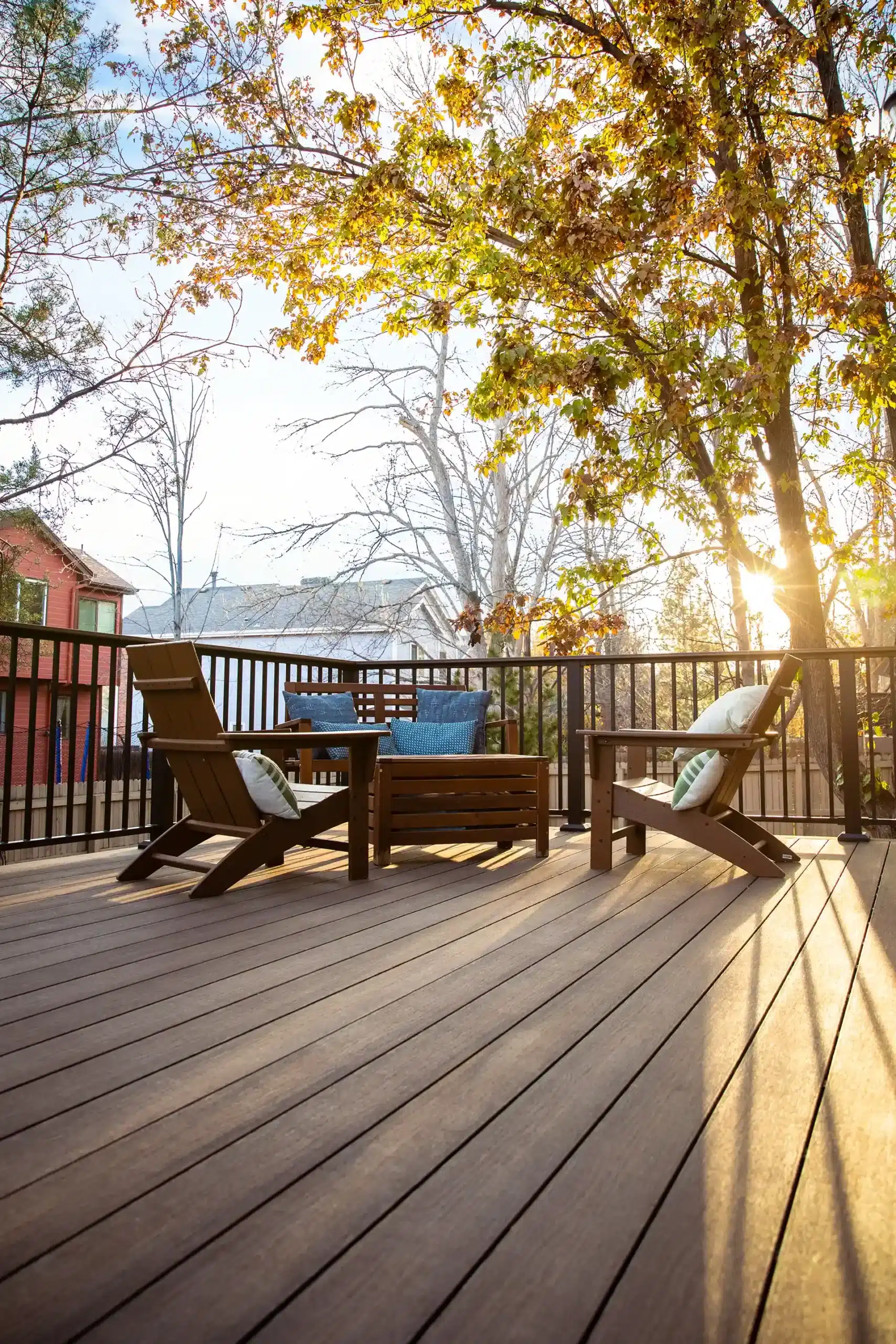 Sunlit composite deck with wooden lounge chairs and a small table, surrounded by trees. 