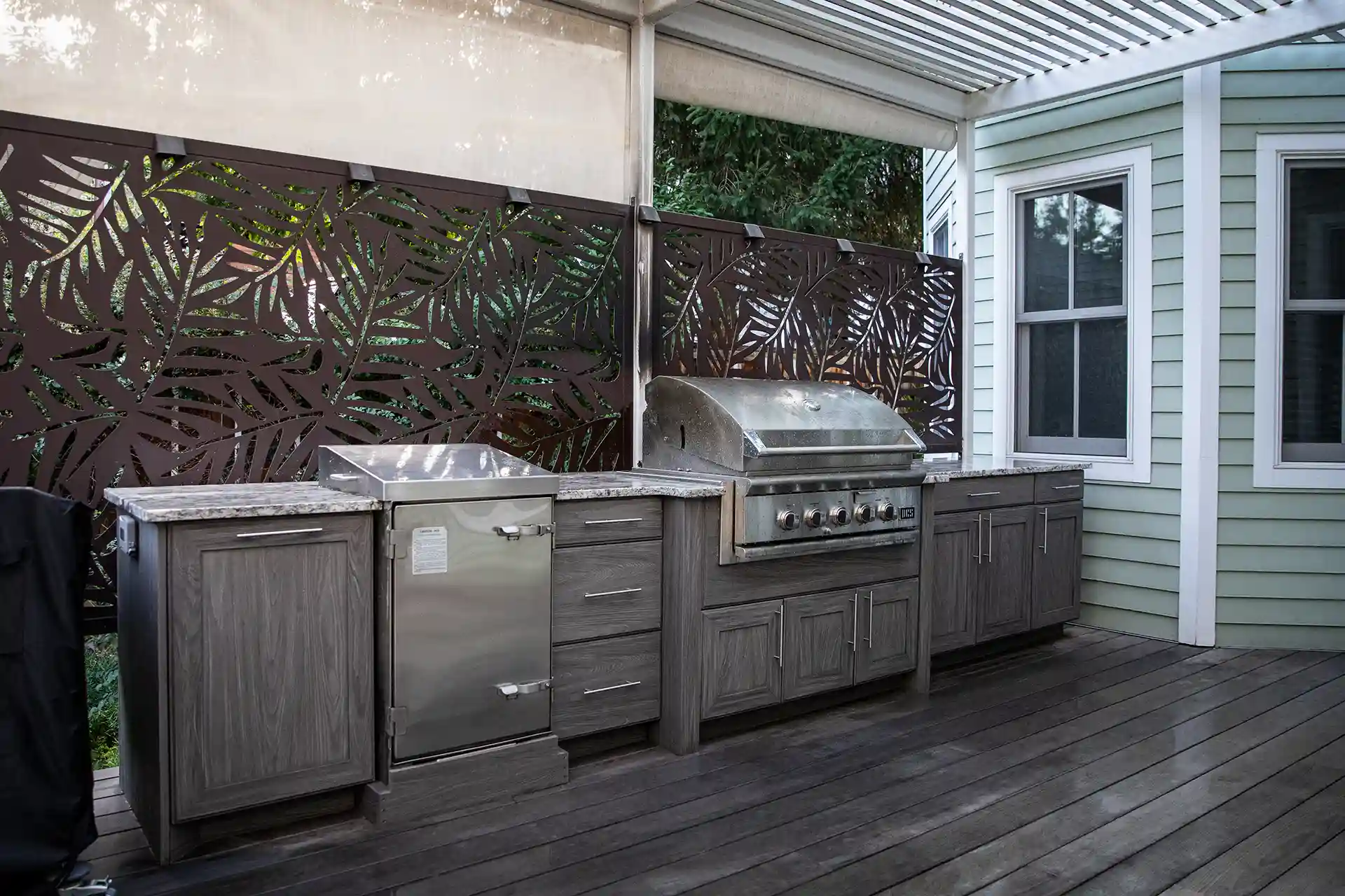 Outdoor kitchen with stainless steel grill, refrigerator, and grey wooden cabinets on a covered deck. 
