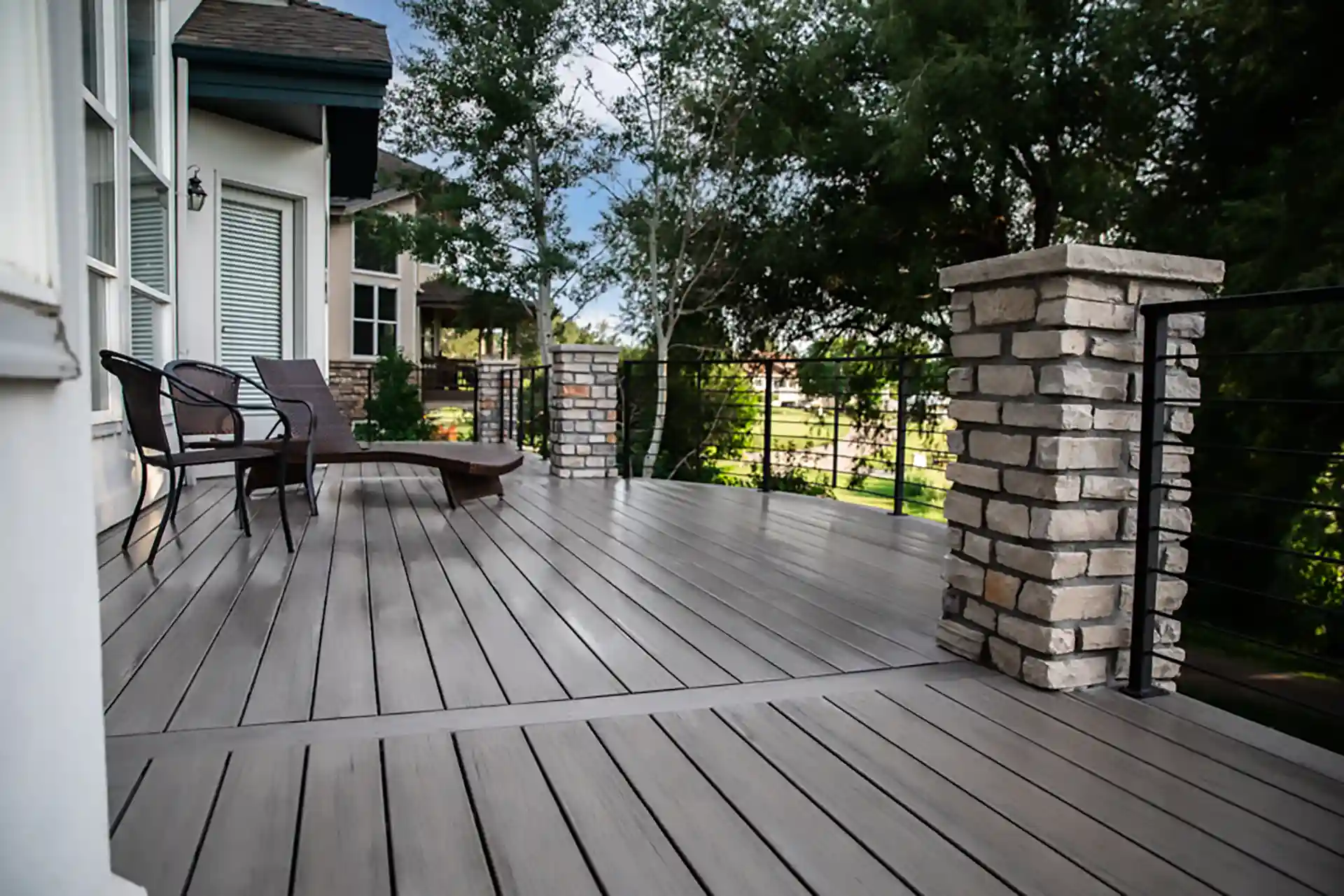 Modern composite deck with cable railing and stone pillars, surrounded by lush greenery. 