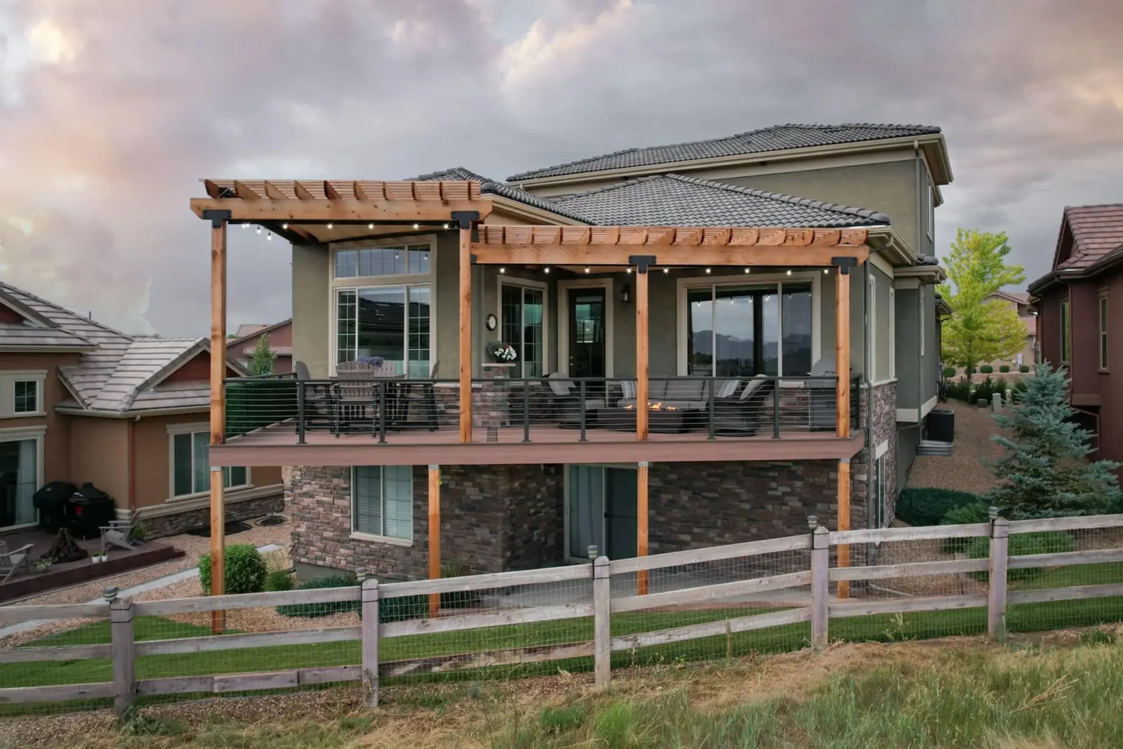 Patio Cover vs Pergola A two-story house with a raised wooden deck and a pergola extending from the second floor. The pergola has an open, slatted roof and is adorned with string lights.