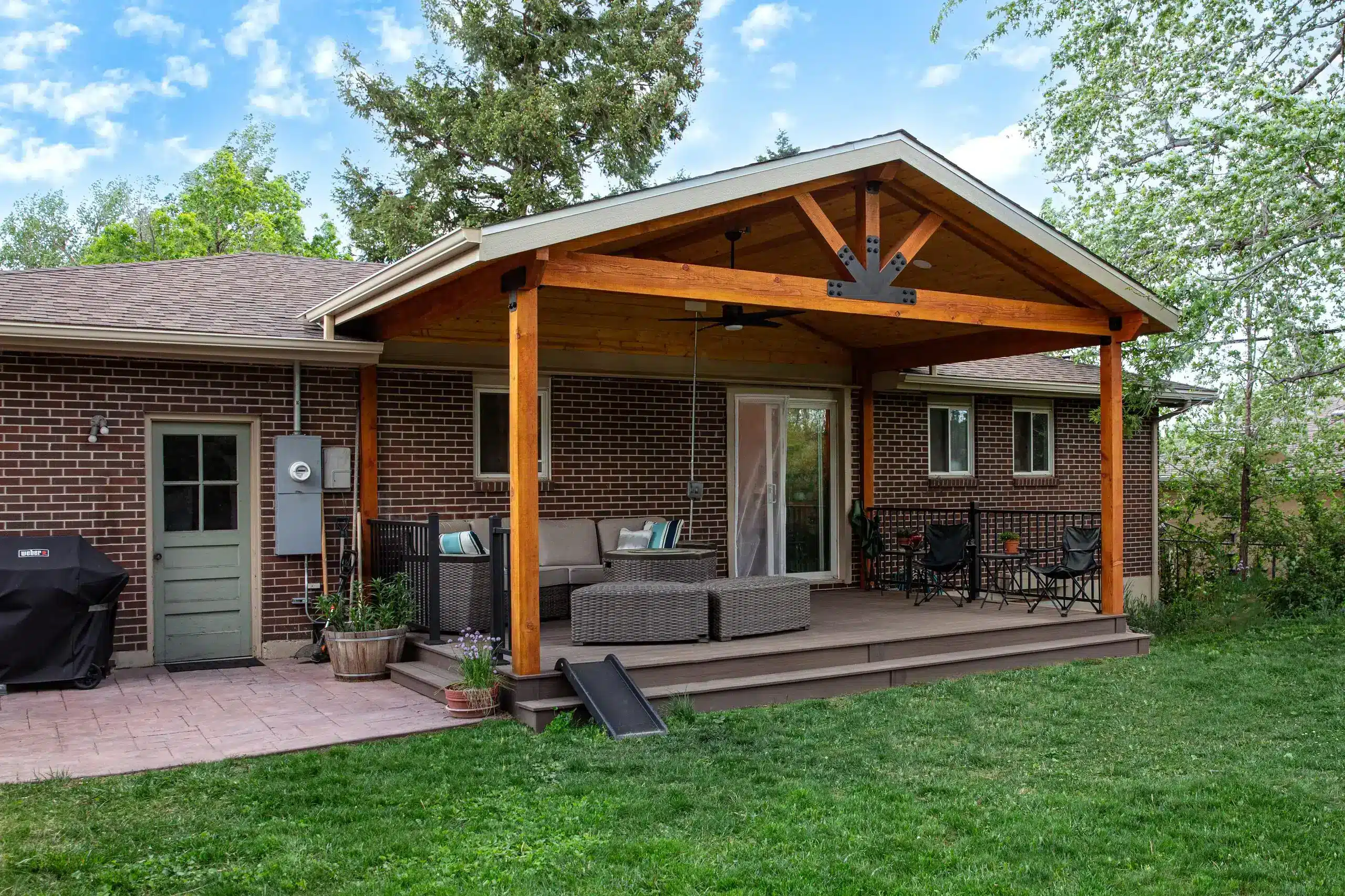 A covered wooden patio attached to a single-story brick house. The patio has a brown composite deck and is supported by large, stained wooden beams and columns. The patio roof is gabled, with exposed wood trusses, and features a dark metal decorative bracket at the peak. A ceiling fan is mounted underneath the roof.