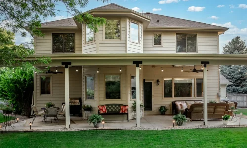 A rear view of a two-story beige house with a covered patio below, furnished with outdoor seating and string lights, overlooking a green lawn.